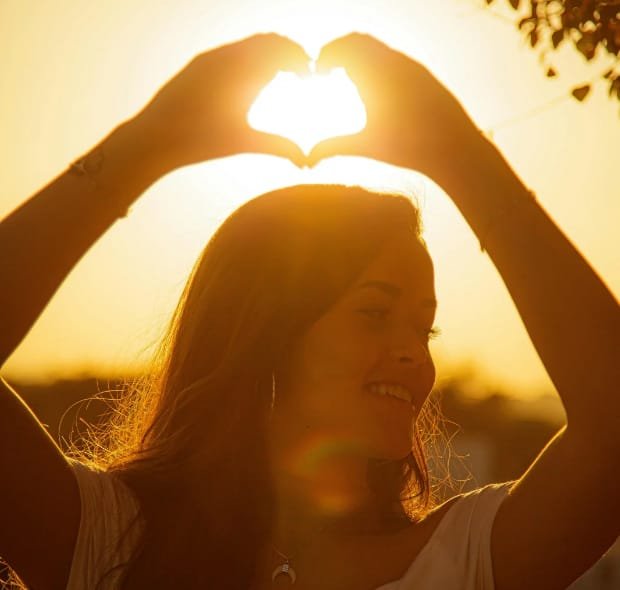 Mulher forma um coração com as mãos diante do pôr do sol, simbolizando amor-próprio e bem-estar emocional.